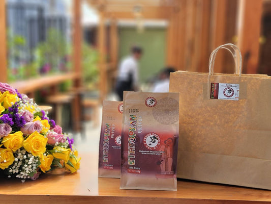Two packages of Ethiopian coffee and a brown paper bag on a wooden surface with flowers in the background.